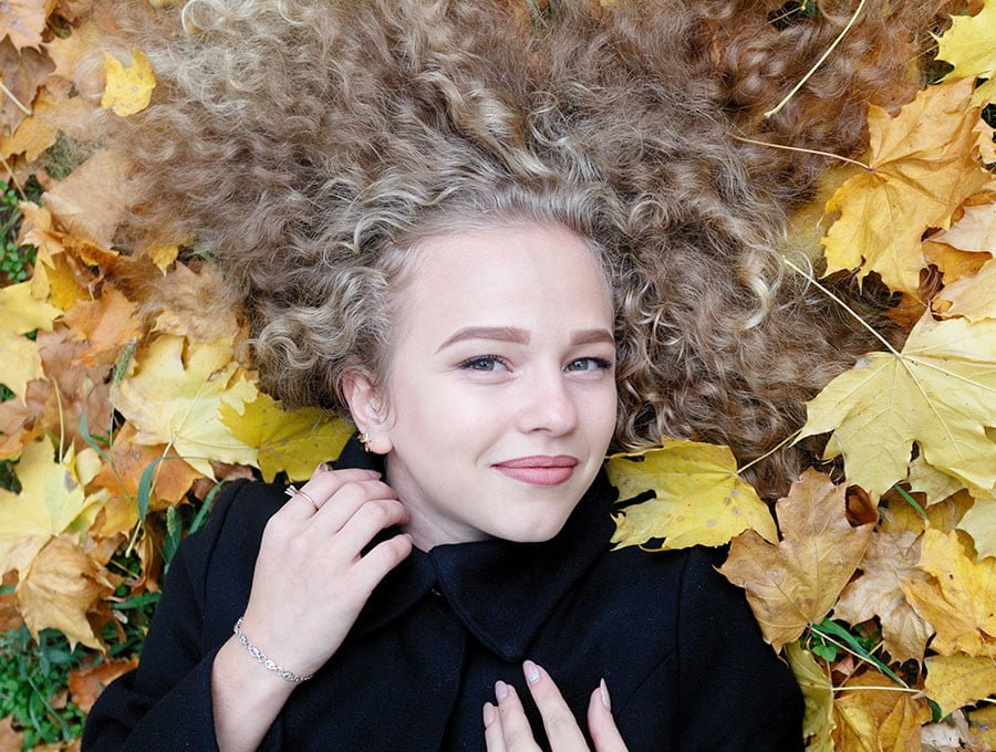 Chica con cabello rizado tumbada sobre las hojas secas del jardín.